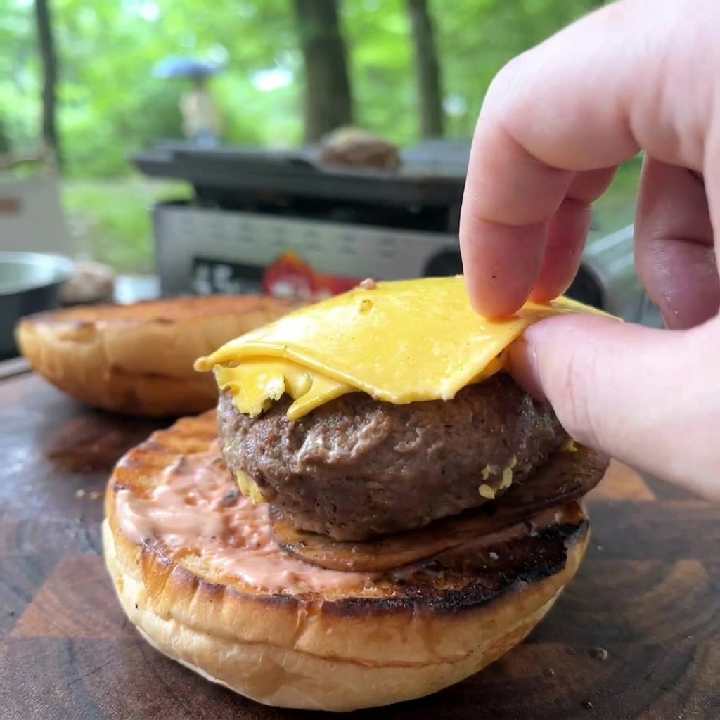Eine Hand legt ein gebratenes Beef-Patty mit geschmolzenem gelben Käse auf ein mit cremiger Sauce bestrichenes Burger-Bun.