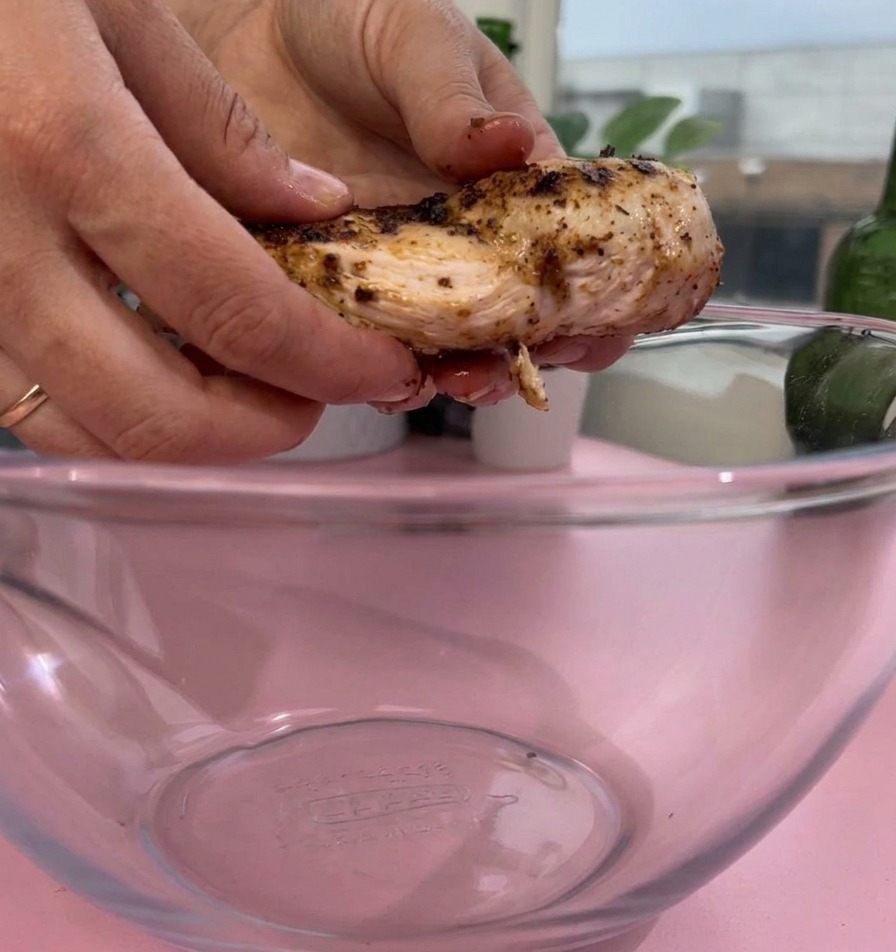 Zwei Hände legen ein Stück gegarte, gewürzte Hähnchenbrust in eine klare Rührschüssel aus Glas auf einer rosa Oberfläche.