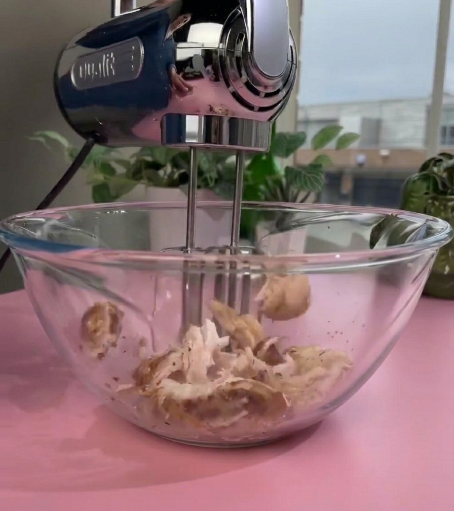 Ein elektrischer Handmixer mit rotierenden Rührbesen zerkleinert gegartes Hähnchenfleisch in einer Glasschüssel zu einer feinen Textur.