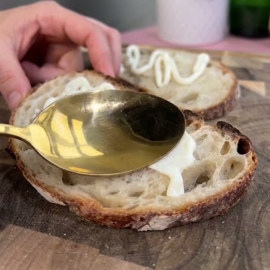 Ein goldener Löffel verstreicht dicke weiße Mayonnaise auf einer gerösteten Scheibe rustikalem Sauerteigbrot.