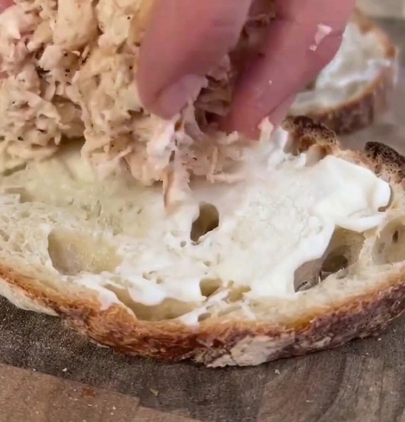 Eine Hand setzt eine großzügige Menge gezupftes, gegartes Hähnchenfleisch auf eine mit Mayonnaise bestrichene Scheibe Sauerteigbrot.
