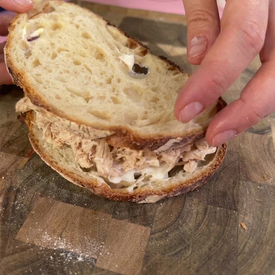 Hände legen eine geröstete Scheibe Sauerteigbrot auf einen Haufen gezupftes Hähnchen, um das Sandwich zu schließen.
