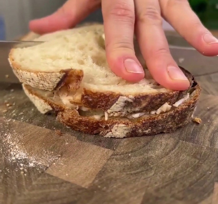 Eine Hand drückt flach auf ein großes Hähnchen-Sauerteig-Sandwich auf einem Holzbrett, während ein Messer zum Anschneiden bereitgesetzt wird.