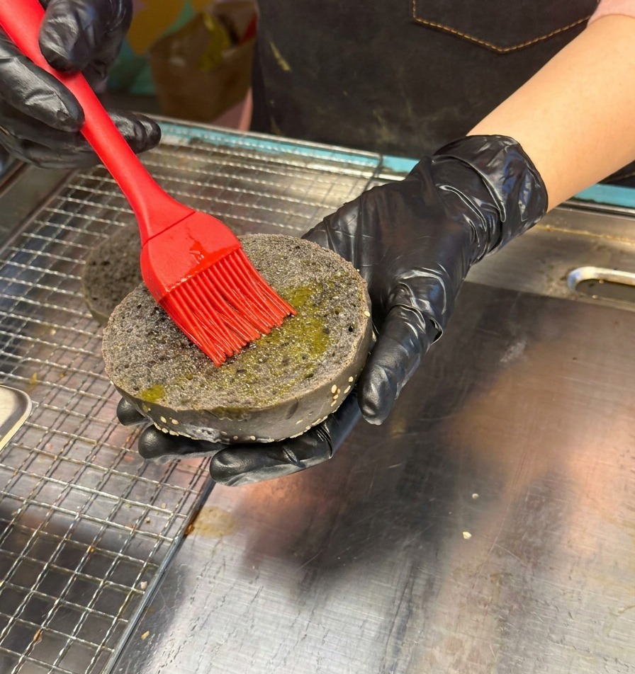 Eine Hand in einem schwarzen Handschuh bestreicht die Innenseite eines schwarzen Sesam-Burger-Buns mit einem roten Silikonpinsel mit Öl.