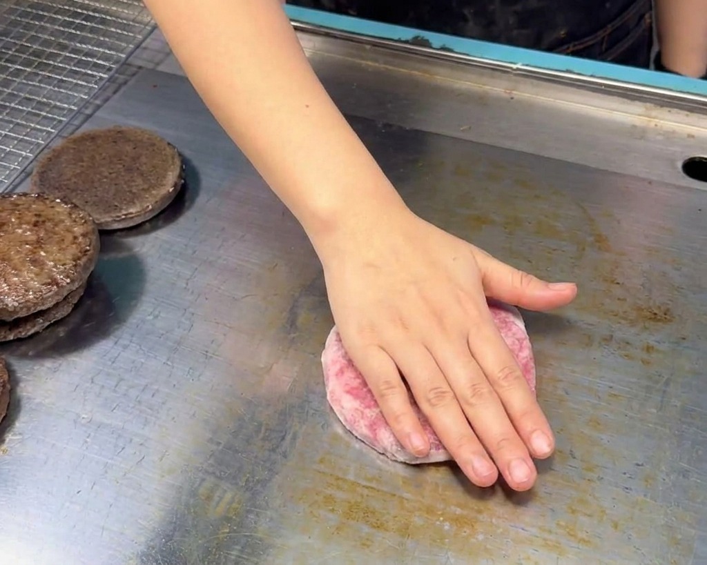 Eine Hand legt ein rohes, rosa Rindfleisch-Patty auf eine saubere Edelstahl-Grillplatte neben getoastete Brötchenhälften.