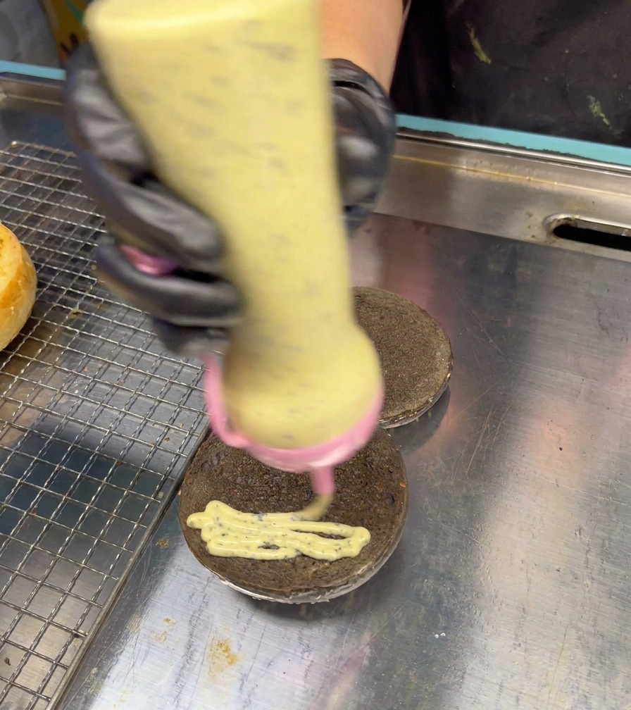 Eine Dosierflasche trägt eine cremige, gesprenkelte Trüffelsauce in Schlangenlinien auf ein schwarzes Burger-Bun auf.