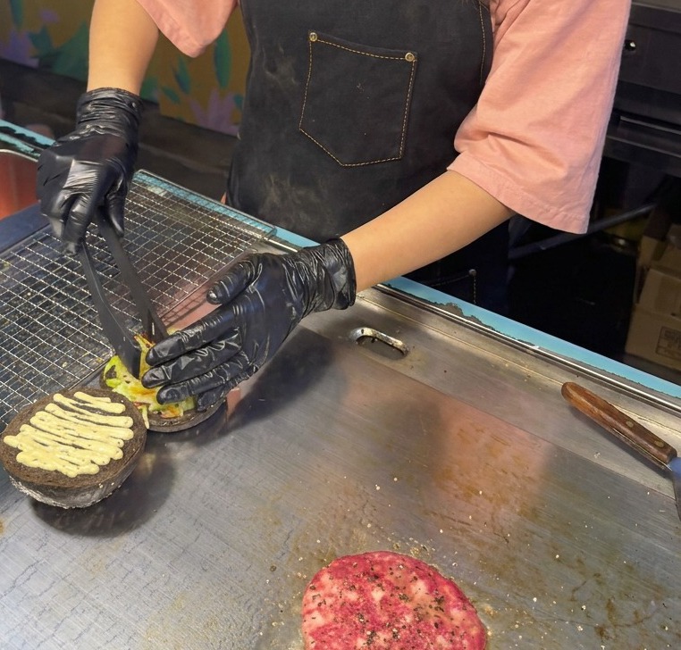 Eine Grillzange legt eine Mischung aus geschnittenem Blattsalat und Rotkohl auf ein bereits mit Trüffelsauce bestrichenes schwarzes Bun.