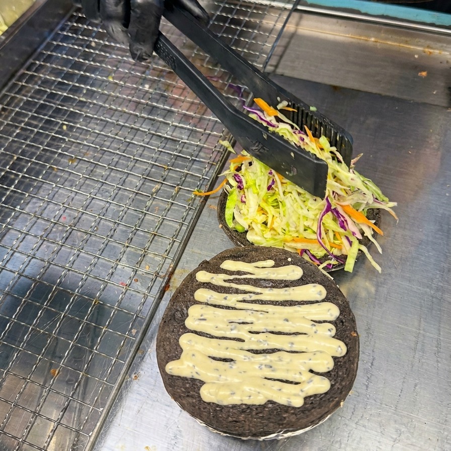 Eine Person legt vorsichtig mit einer Zange eine leuchtend rote Tomatenscheibe auf das Salatbett eines schwarzen Aktivkohle-Burger-Buns.