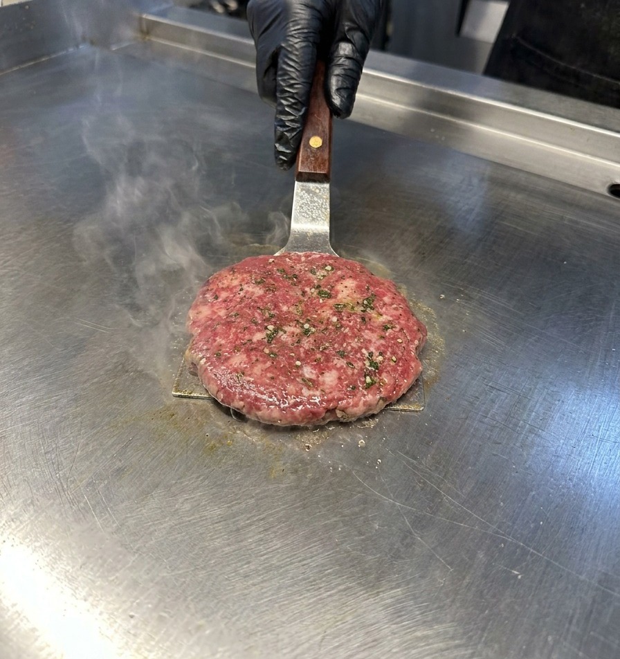 Ein Metallwender drückt leicht auf ein Rindfleisch-Patty auf einer Edelstahl-Grillplatte, während Dampf aufsteigt.