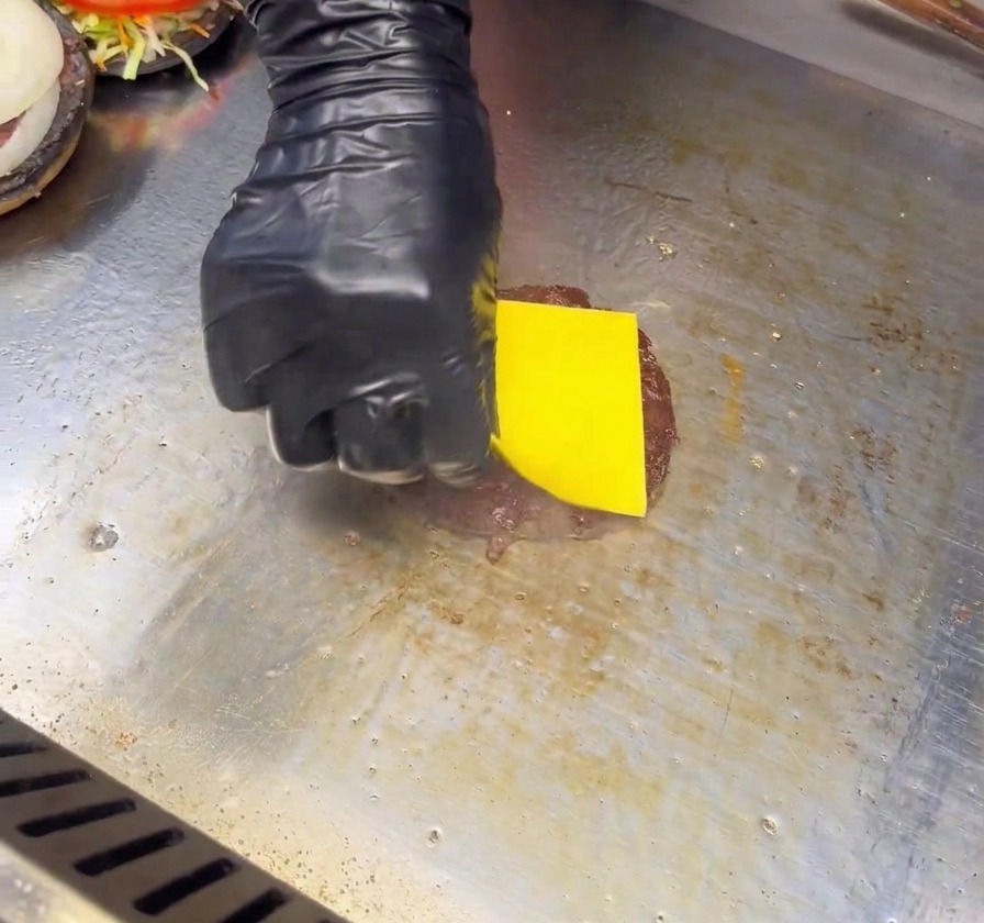 Eine Hand im schwarzen Handschuh legt eine Scheibe gelben Käse auf ein angebratenes Burger-Patty auf einer Grillplatte.