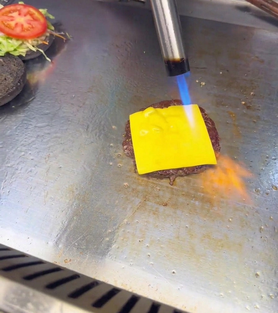 Eine blaue und orangefarbene Flamme eines Küchenbrenners bringt eine Scheibe Käse auf einem Burger-Patty zum Schmelzen.