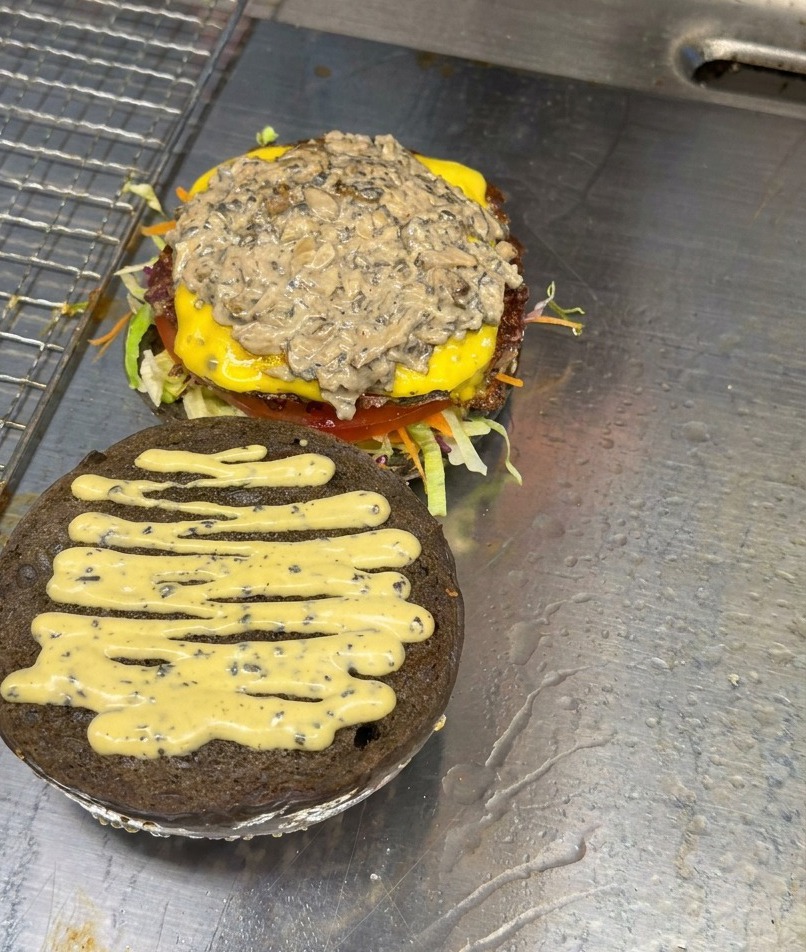 Eine Schicht in Scheiben geschnittener, gebratener Pilze liegt auf einem fertig mit Käse und Sauce geschichteten Burger-Patty.