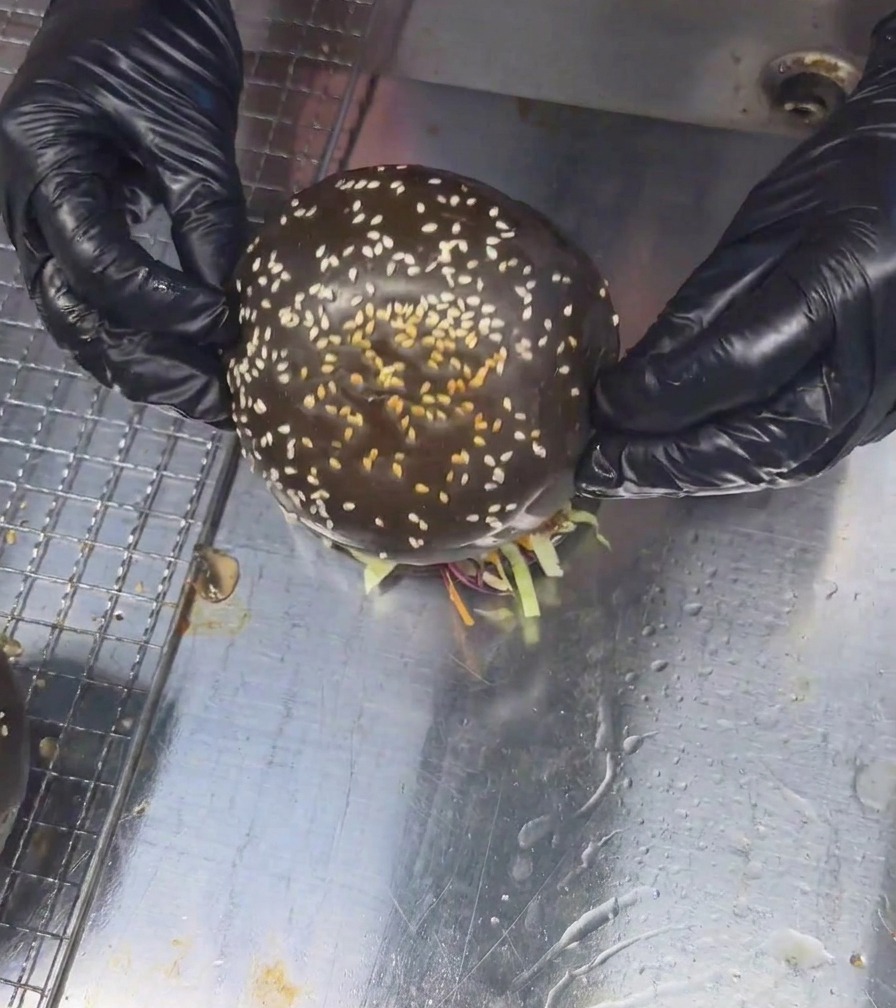 Zwei behandschuhte Hände setzen ein schwarzes Sesam-Brötchen auf einen hohen, fertig geschichteten Pilz-Beef-Burger.
