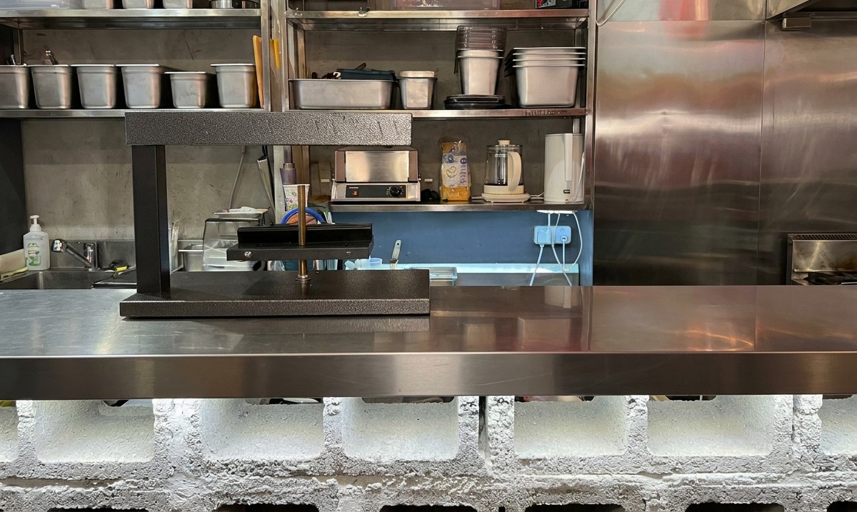 Ein schwarzer elektrischer Raclette-Ofen steht einsatzbereit auf einer Arbeitsfläche aus Edelstahl.