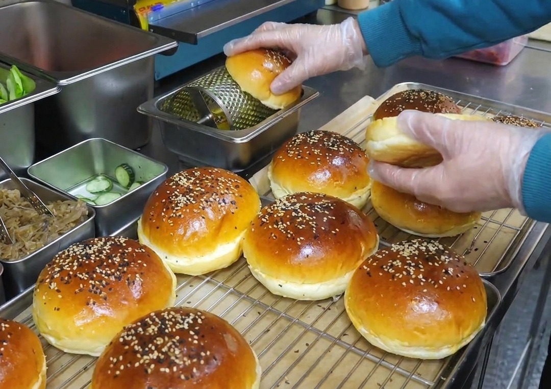 Hände in Plastikhandschuhen rollen Burger-Brötchen über eine rotierende Walze zum Auftragen von geschmolzener Butter.