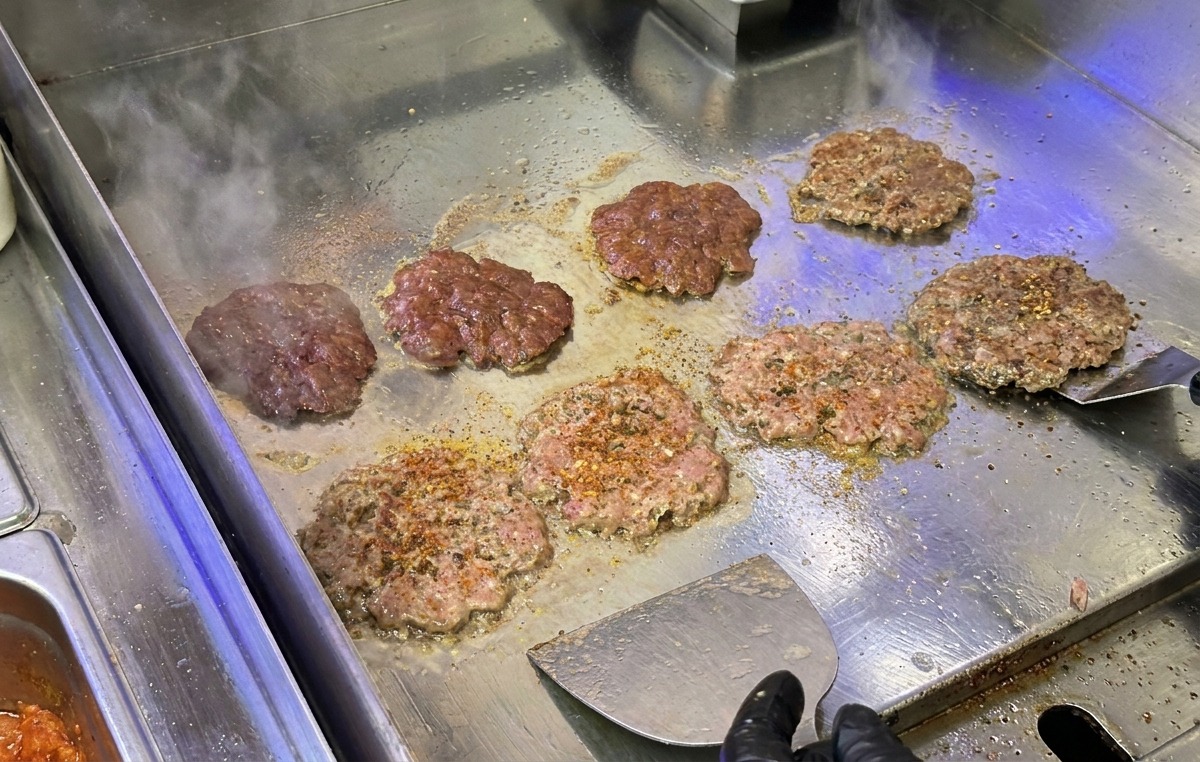 Rinder-Patties werden auf einer Edelstahl-Grillplatte mit einem Fleischwender fest angedrückt und scharf angebraten.