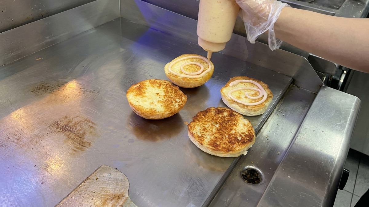 Eine cremige, rosafarbene Sauce wird in kreisenden Bewegungen auf getoastete Brioche-Brötchen auf einer Grillplatte aufgetragen.