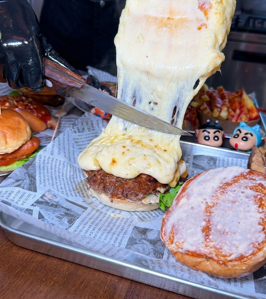 Ein großes Messer schabt eine dicke Schicht geschmolzenen, schäumenden Käse von einem Laib auf ein Rinder-Patty.