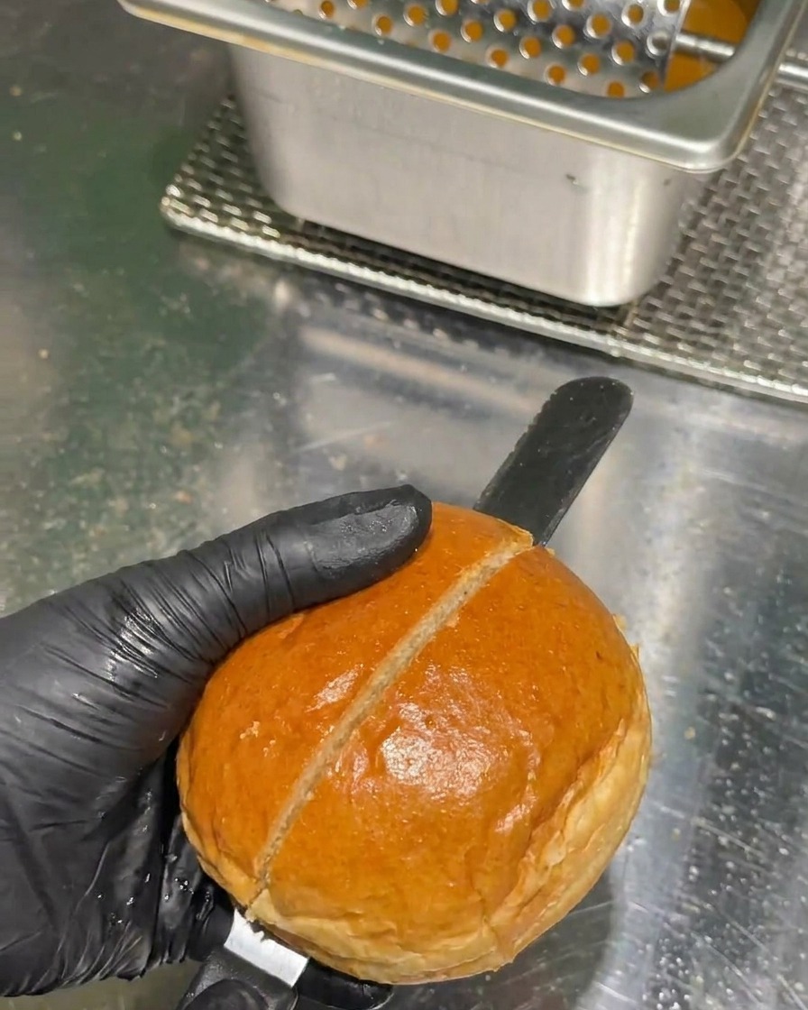 Eine Hand in einem schwarzen Handschuh schneidet ein goldbraunes Burgerbrötchen mit einem langen Sägemesser auf einer Edelstahl-Arbeitsplatte in zwei Hälften.