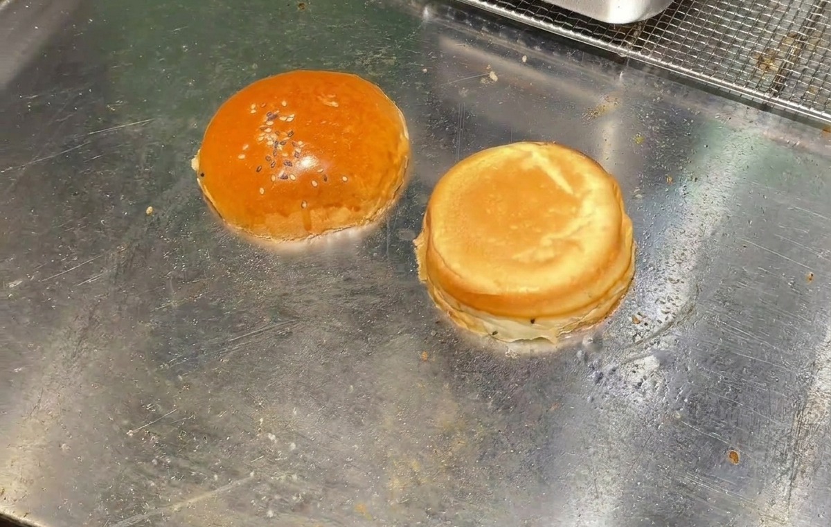 Zwei Hälften eines Burgerbrötchens werden mit der Schnittseite nach unten auf einer flachen Grillplatte getoastet.
