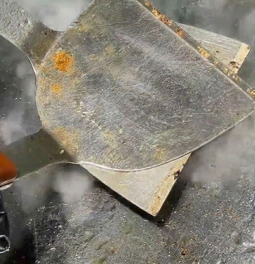Ein Metallspatel drückt ein rundes Fleischbällchen auf einem heißen Grill zu einem flachen Patty, während Dampf aufsteigt.