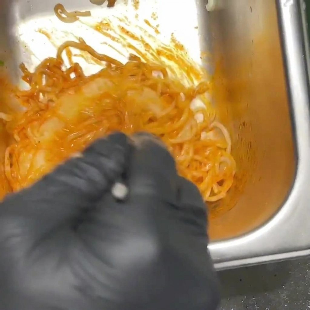 Scharfe, orangefarbene Ramen-Nudeln werden von einer Person mit schwarzen Handschuhen in einem Metallbehälter vermischt.