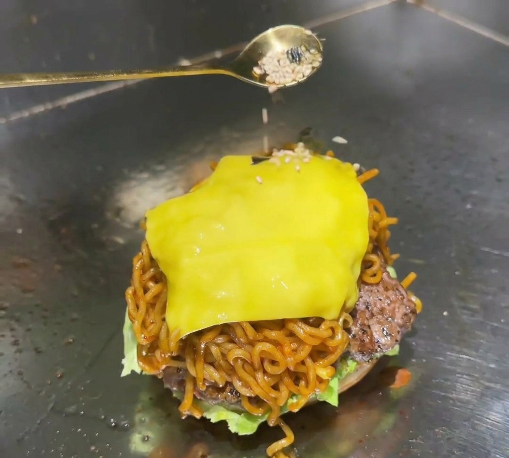 Ein goldener Löffel streut eine Mischung aus weißem Sesam und schwarzen Algenflocken auf den geschmolzenen Käse eines Ramen-Burgers.