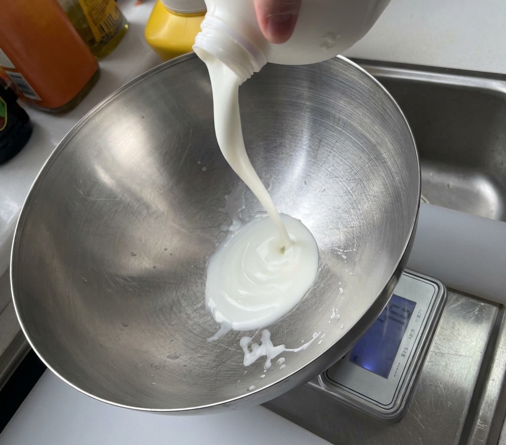 Weißer Joghurt wird aus einem Plastikbehälter in eine große Rührschüssel aus gebürstetem Metall gegossen.