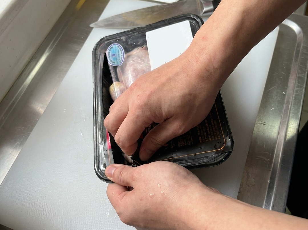 Hände öffnen eine in Plastik eingepackte Schale mit rohen Hähnchenschenkeln auf einem weißen Schneidebrett neben einem Küchenmesser.