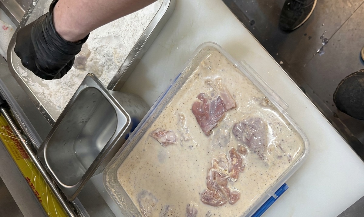 Behandschuhte Hände legen mariniertes Hähnchenfleisch in eine Schale mit gewürztem Paniermehl.