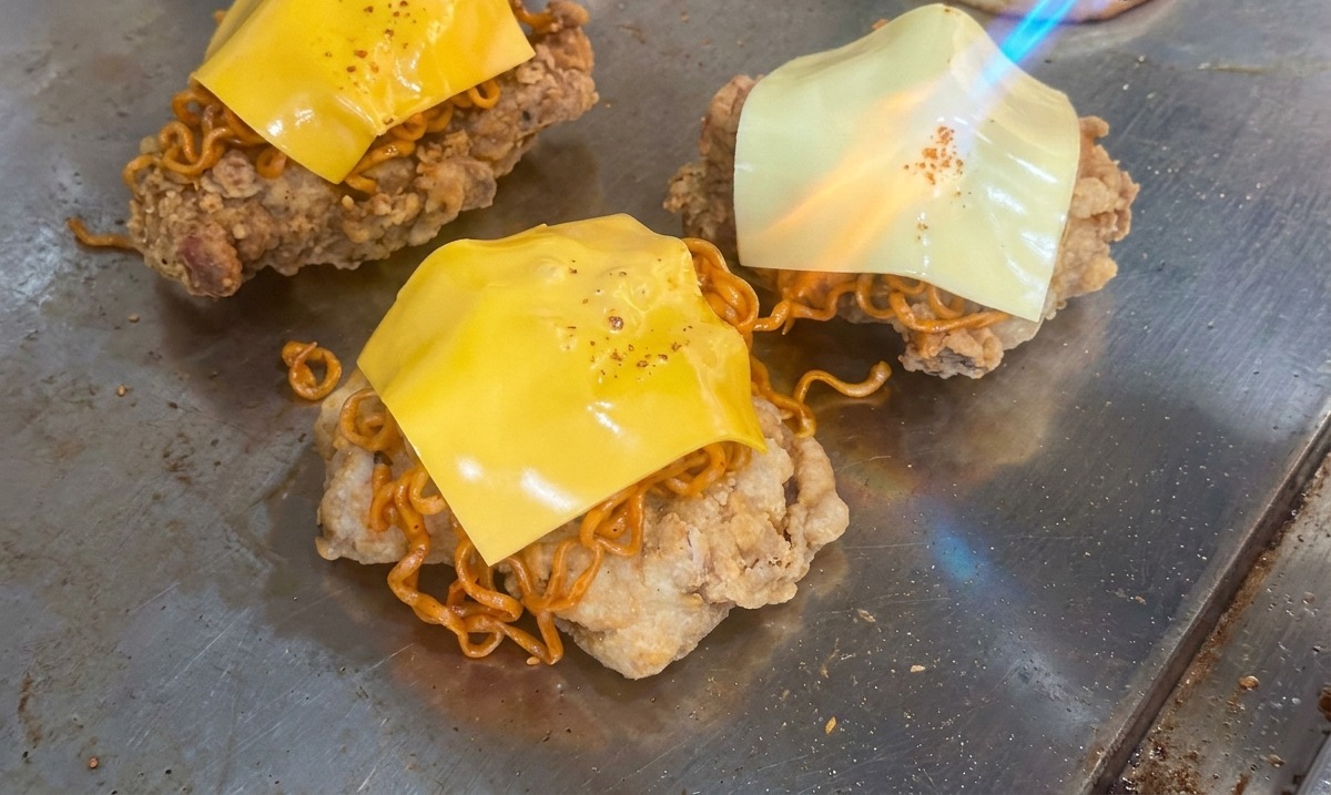 Die Flamme eines Bunsenbrenners schmilzt ein Stück gelben Käse über einem Berg aus scharfen Nudeln und gebratenem Hähnchen auf einem Grill.