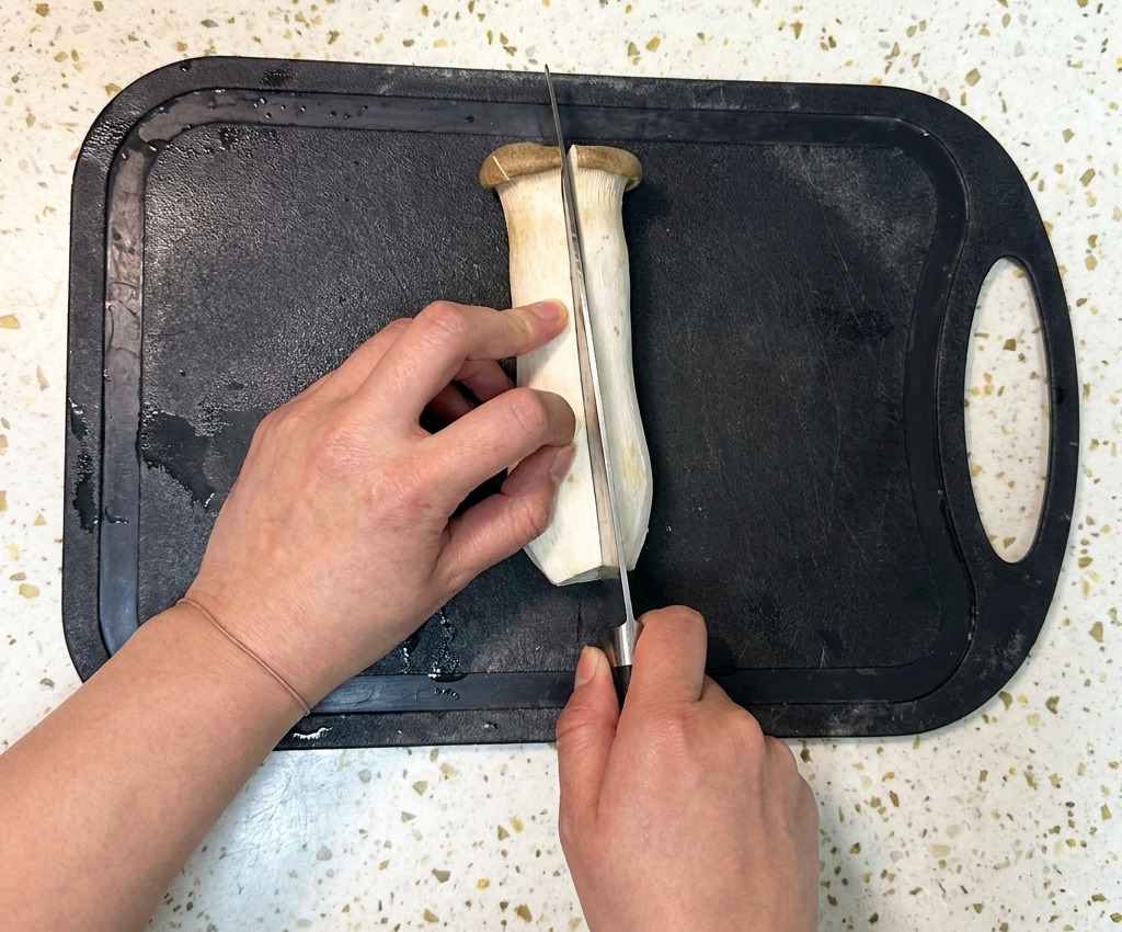 Eine Person schneidet einen großen Kräuterseitling mit einem Kochmesser auf einem schwarzen Schneidebrett der Länge nach durch.