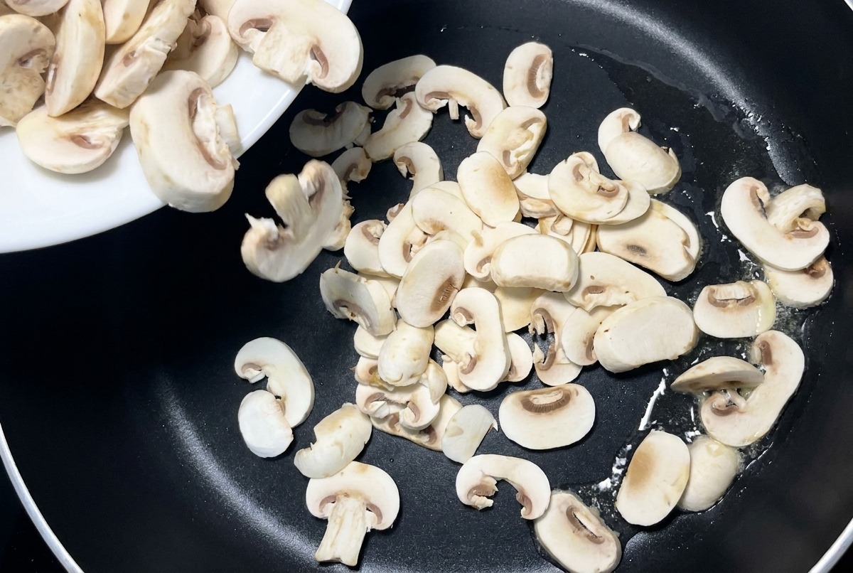 Geschnittene weiße Pilze werden von einem weißen Keramikteller in eine schwarze beschichtete Pfanne gegeben.