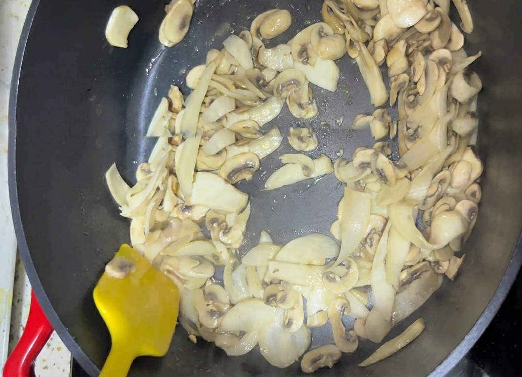 Geschnittene Pilze werden in einer dunklen Pfanne mit einem leuchtend gelben Pfannenwender umgerührt.