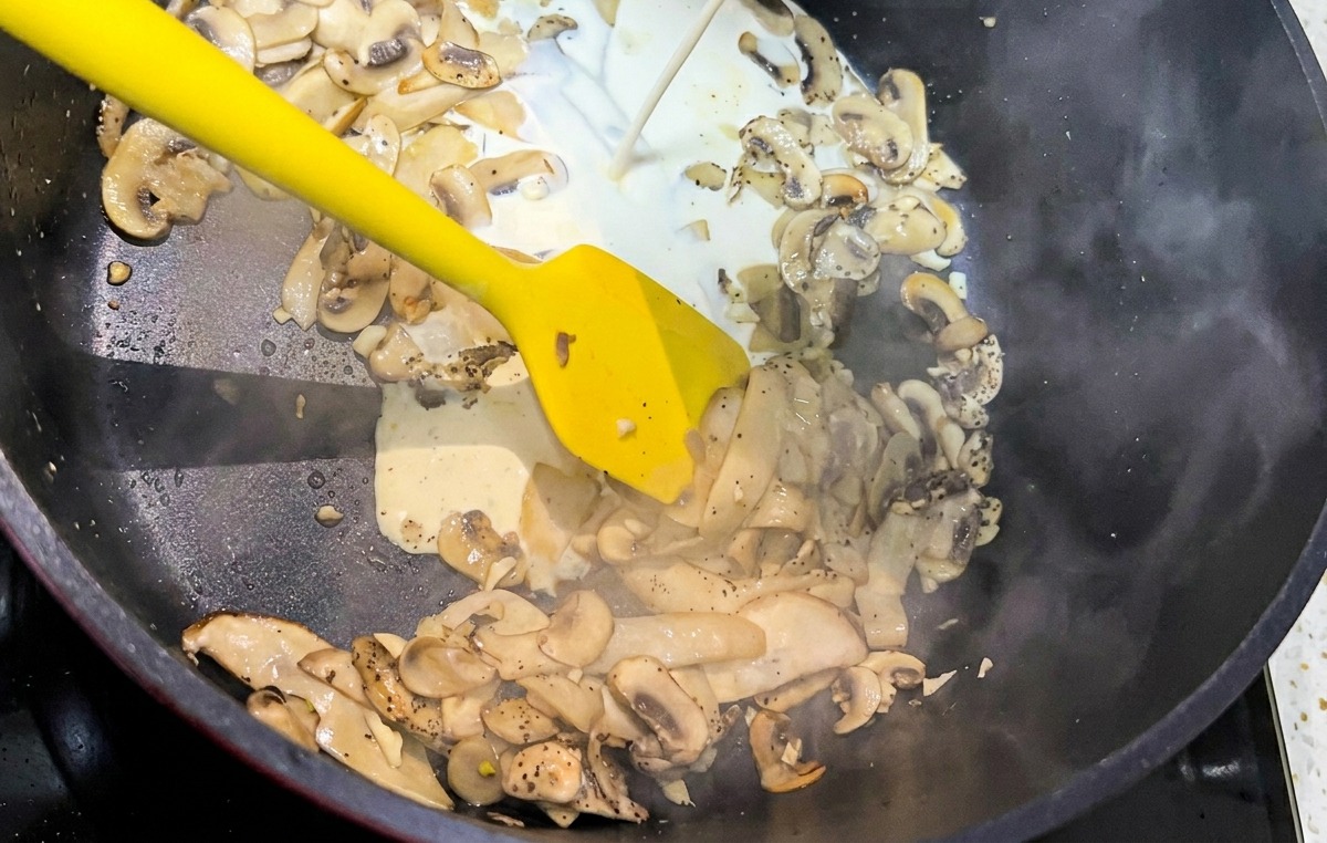 Ein Schuss weiße Schlagsahne wird in eine schwarze Pfanne voller gebratener Pilzscheiben gegossen.