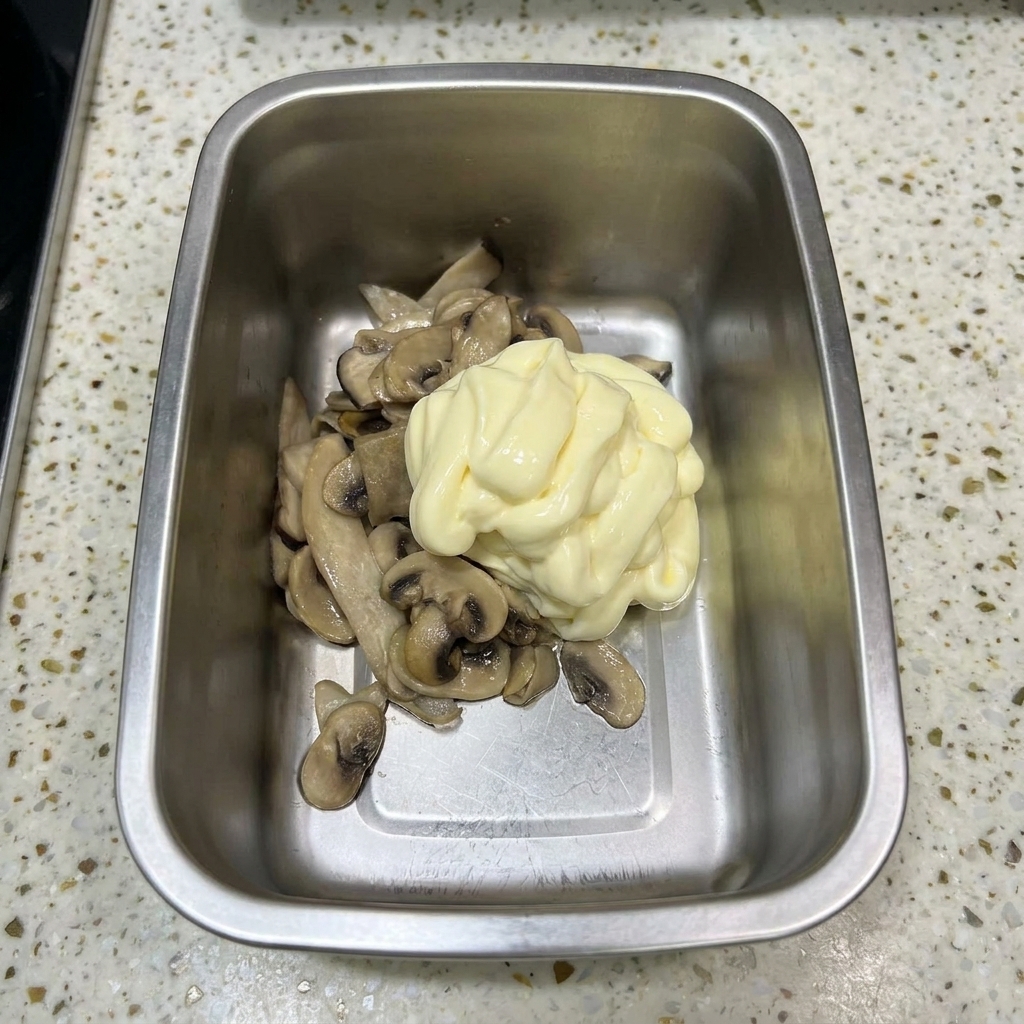 Ein Metallbehälter mit gebratenen Pilzen, gekrönt von einem großen Klecks blassgelber Mayonnaise.