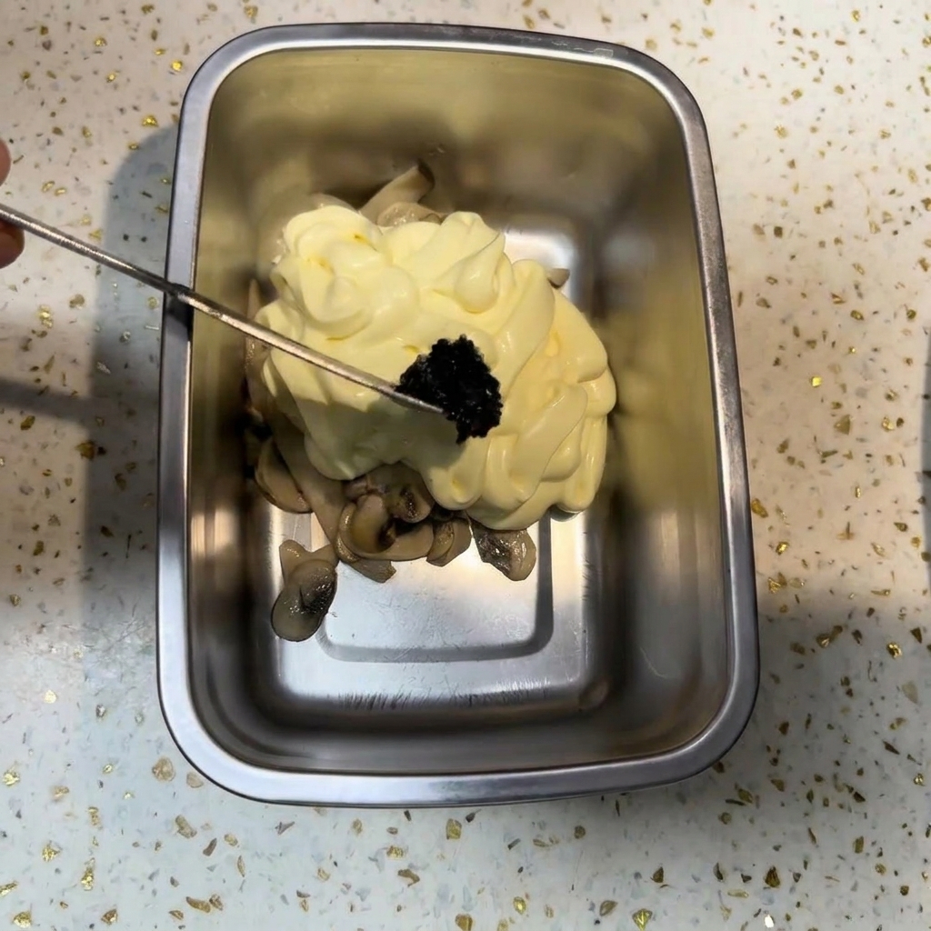 Ein kleines Metallwerkzeug gibt eine Portion dunkle schwarze Trüffelpaste auf einen Berg aus Mayonnaise und Pilzen.
