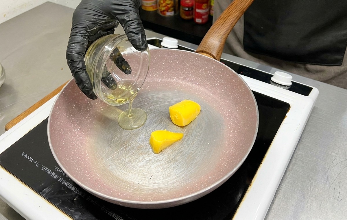 Zwei Stücke gelbe Butter in einer Pfanne, während Olivenöl aus einer Glasschüssel hinzugegossen wird.
