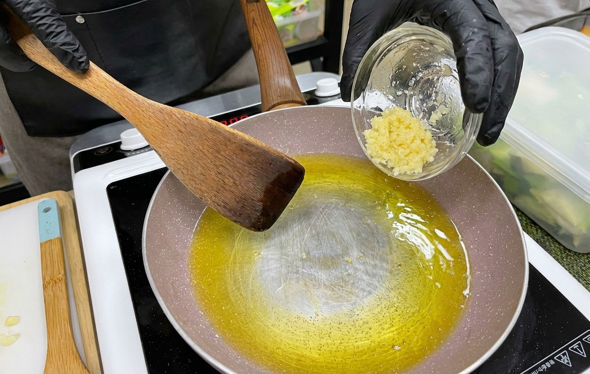 Eine Hand in einem schwarzen Handschuh gibt gehackten Knoblauch aus einer kleinen Schüssel in eine Pfanne mit geschmolzener Butter und Öl.