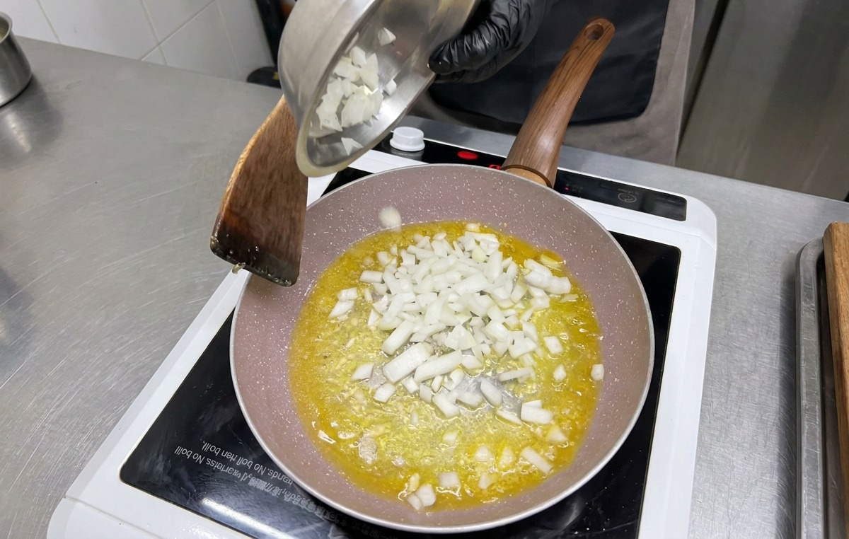 Gewürfelte weiße Zwiebeln werden aus einer Metallschüssel in die Pfanne zu Knoblauch und Butter gegeben.
