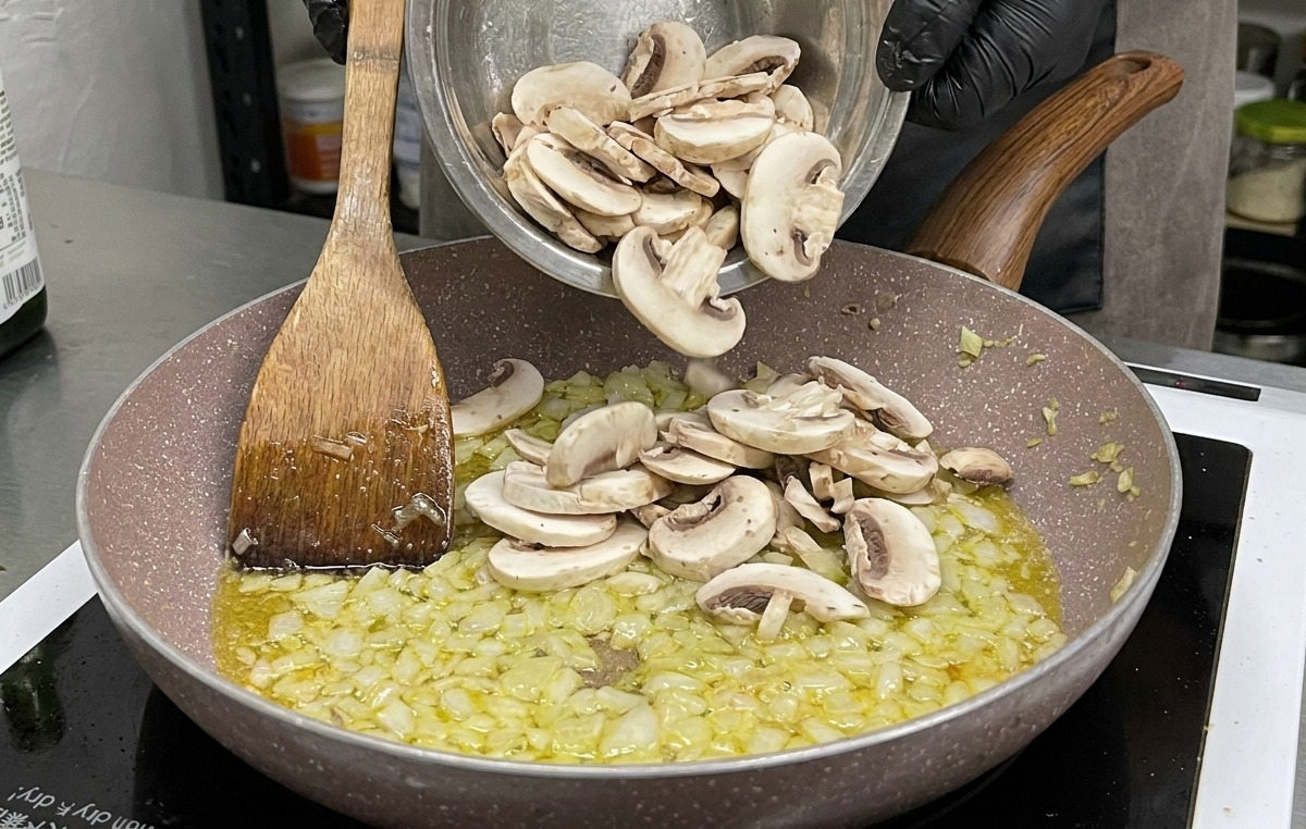 Eine große Schüssel mit rohen, in Scheiben geschnittenen weißen Champignons wird zu den angedünsteten Zwiebeln und dem Knoblauch in die Pfanne gegeben.
