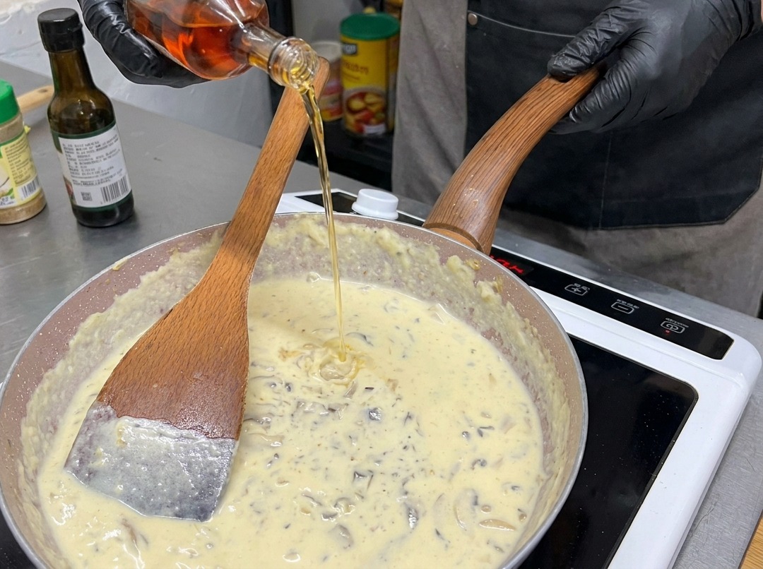 Eine Person mit schwarzen Handschuhen gießt eine bernsteinfarbene Flüssigkeit aus einer Glasflasche in eine Pfanne mit cremiger Pilzsoße.