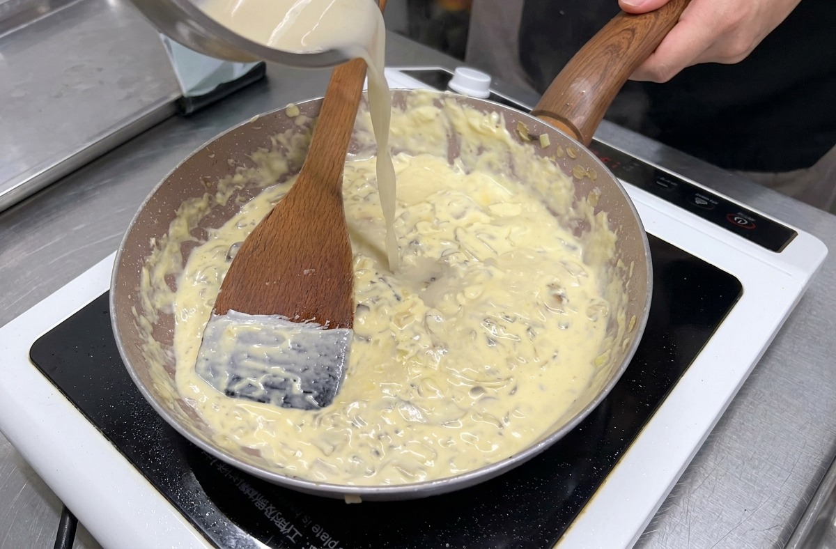 Klare Brühe wird aus einer Edelstahlschüssel in eine Pfanne mit köchelnden Pilzen und Sahne gegossen.