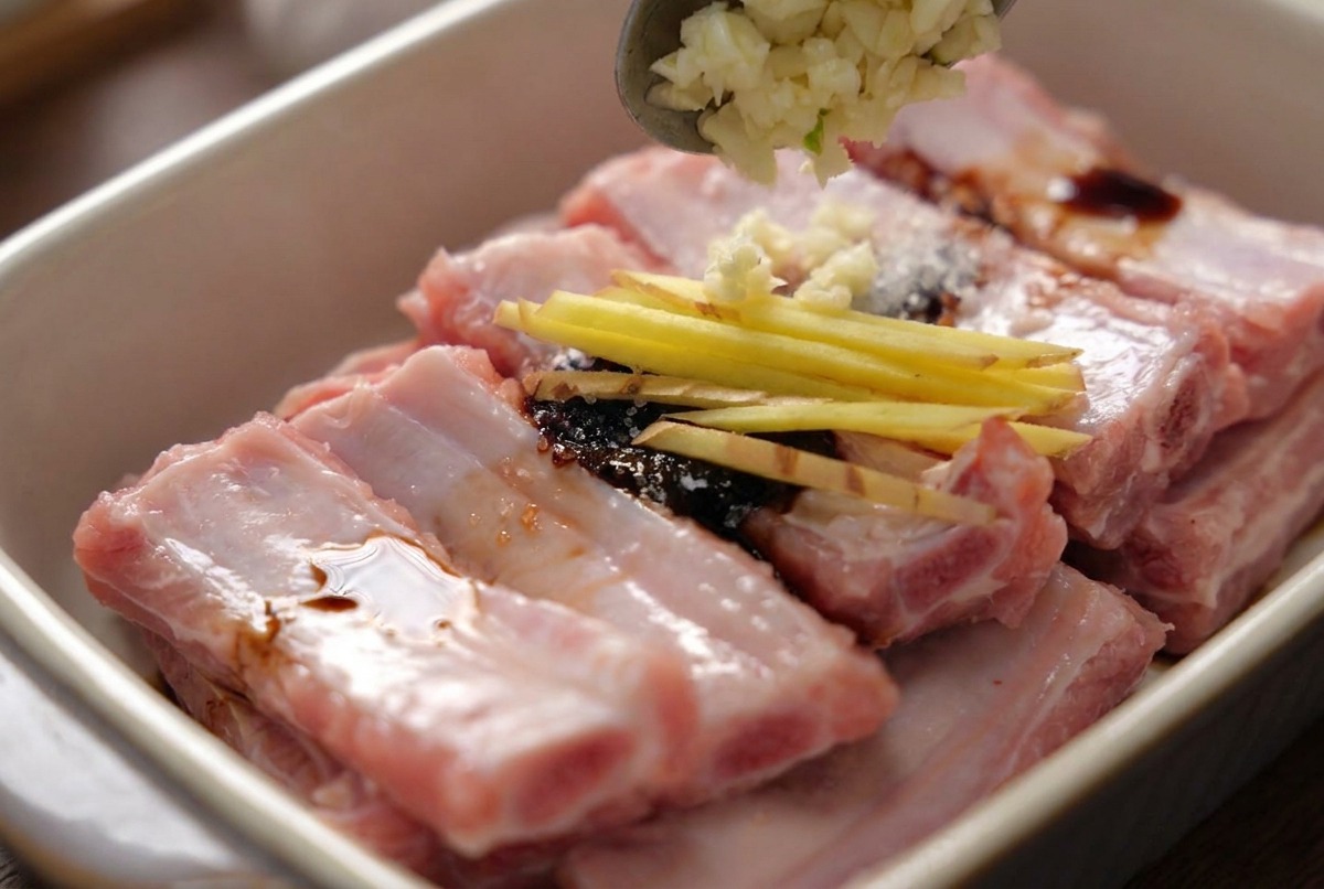 Ein Löffel gibt gehackten Knoblauch auf die gewürzten Schweinerippchen, die bereits mit dünnen Ingwerstreifen garniert sind.