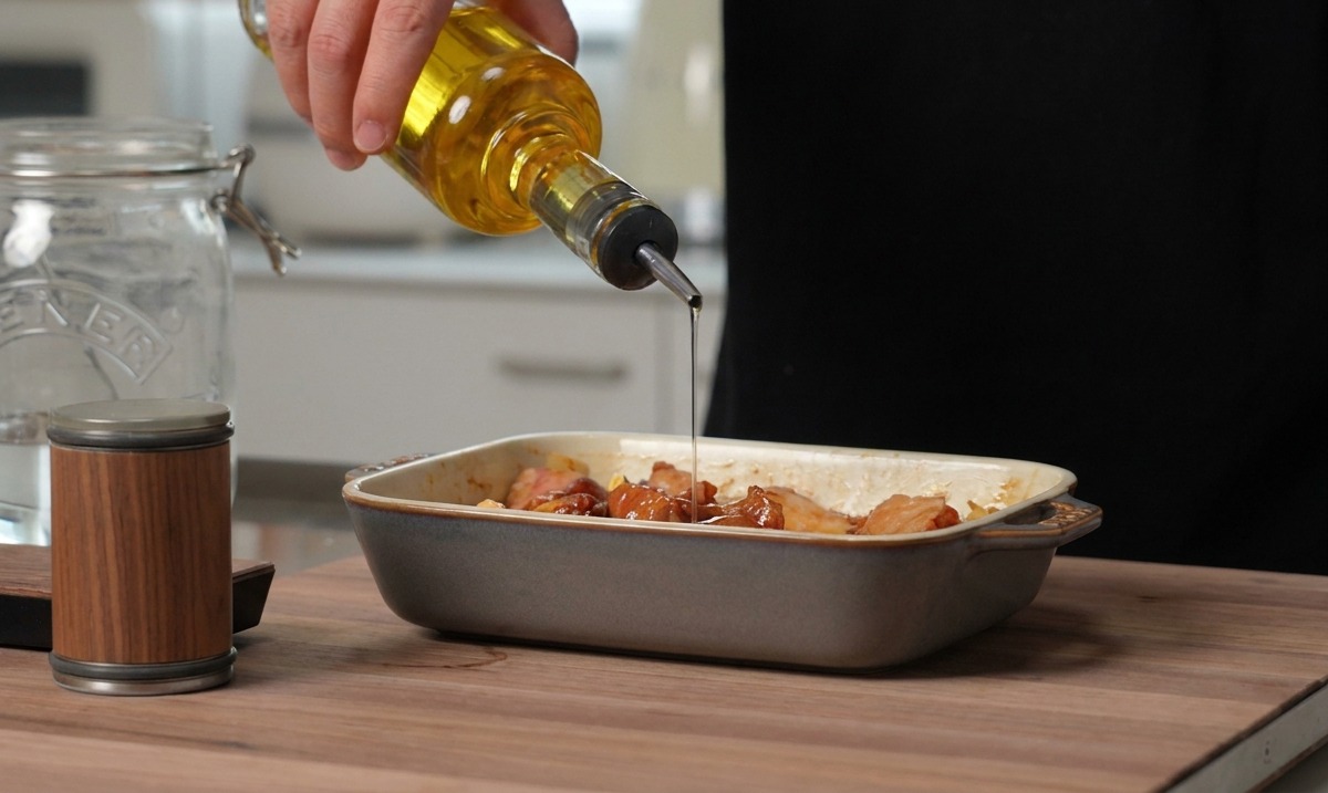 Goldenes Speiseöl wird aus einer Glasflasche auf die gewürzten Schweinerippchen gegossen, die in einer grauen Keramikform liegen.