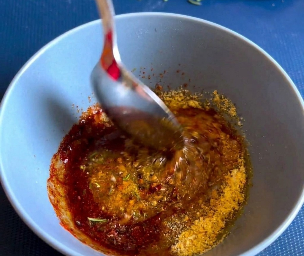 Ein Löffel rührt eine dicke, rotbraune Gewürz- und Kräuterpaste in einer blauen Schale um.