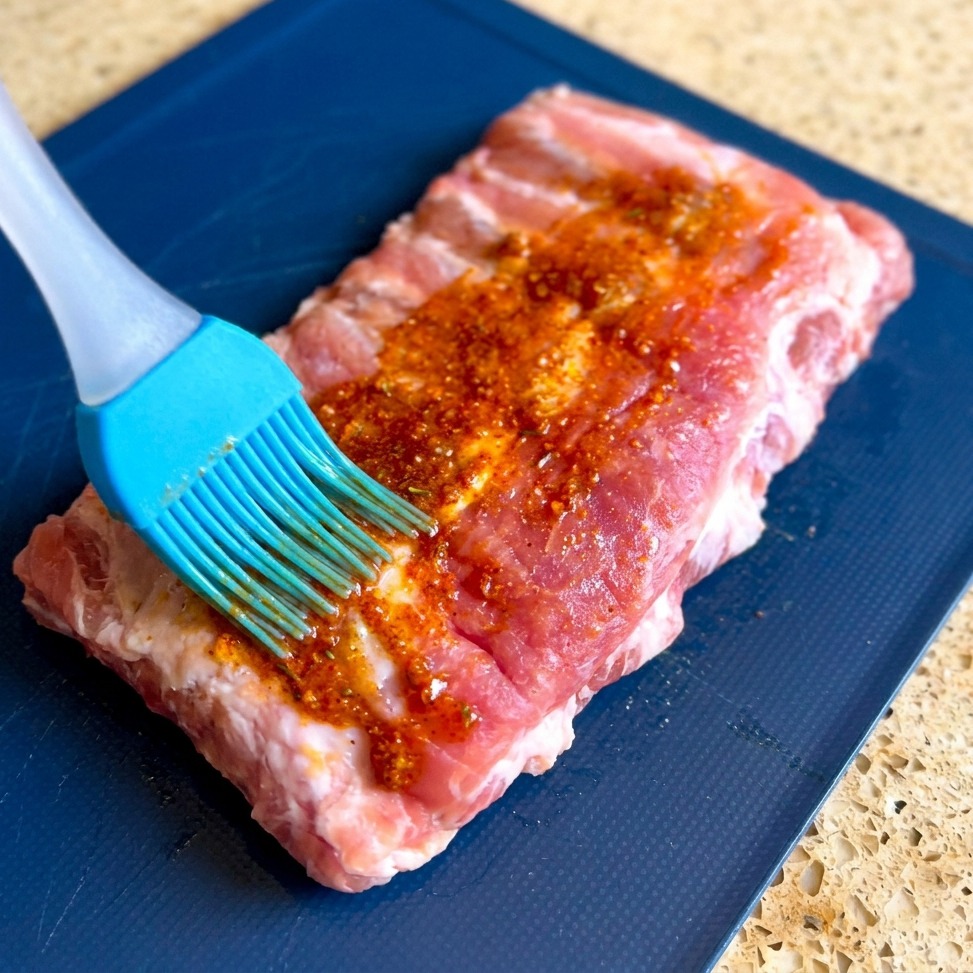Ein blauer Silikonpinsel verstreicht eine dicke, rote Marinade auf einem rohen Stück Schweinerippchen.