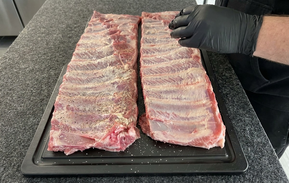 Zwei rohe Schweinerippchen-Racks werden auf einem schwarzen Küchenbrett mit einer Gewürzmischung aus Salz, Pfeffer und Knoblauchpulver bestreut.