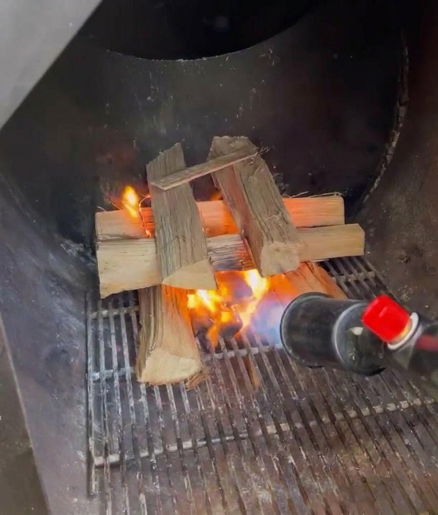 Ein Gasbrenner wird verwendet, um gestapelte Eichenholzscheite in der metallenen Firebox eines Smokers zu entzünden.