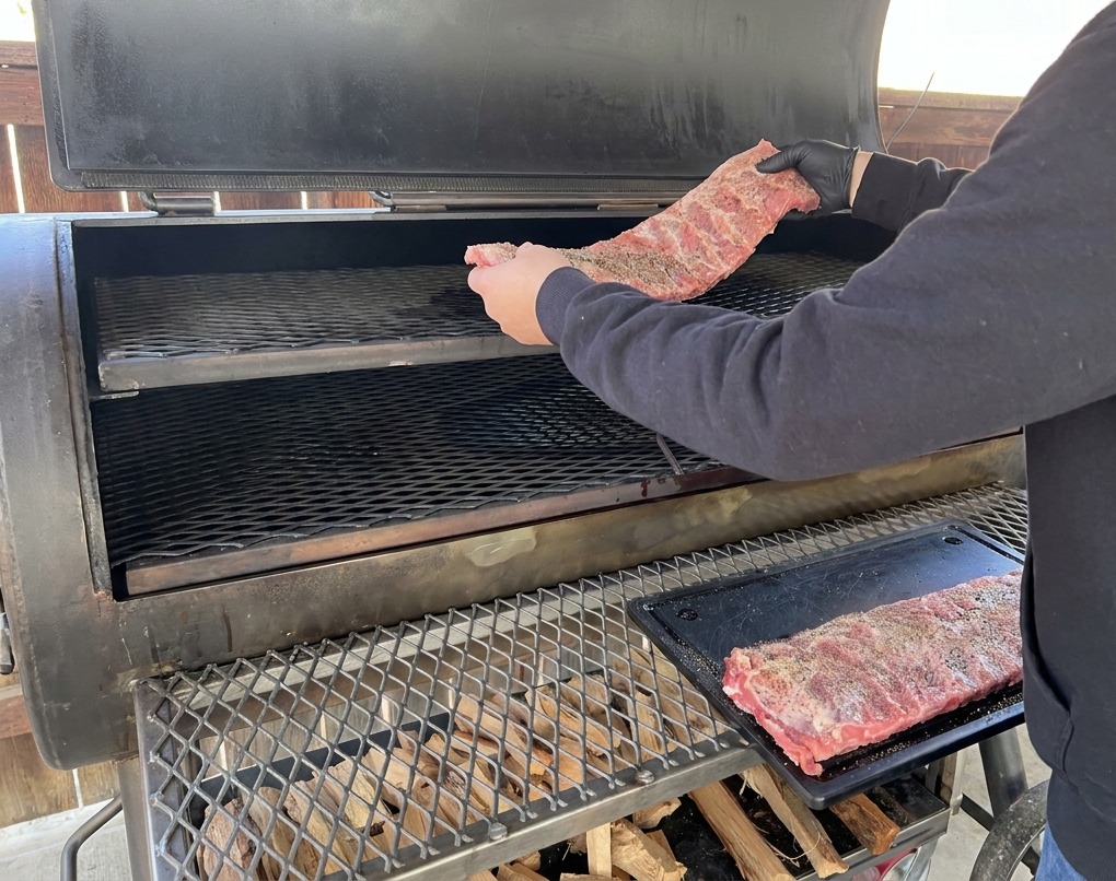Eine Person in einem schwarzen Hoodie und schwarzen Handschuhen legt ein gewürztes Schweinerippchen-Rack auf die obere Schiene eines großen Offset-Smokers.